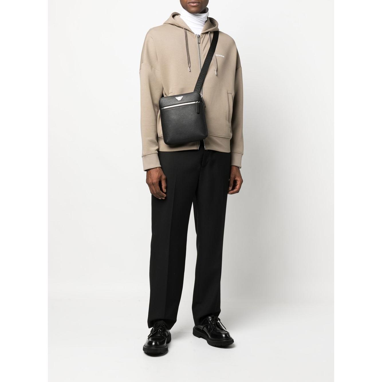 Emporio Armani ASV black faux-leather shoulder bag with eagle plate, worn crossbody by model in beige hoodie and black pants.